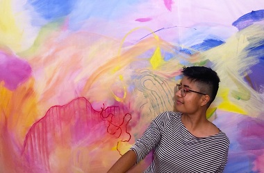 Photo of Katia wearing a black and white striped shirt, glasses and short dark hair in front of a pink, yellow and blue abstract painting.