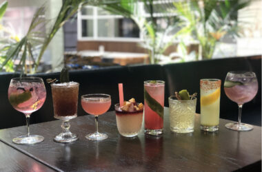 Eight colorful cocktails in varying glasses lined up on a dark table with plants in the background.