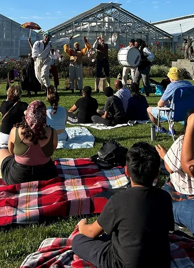 Visitors sit on blankets on a lawn watching a live music performance in front of a greenhouse on a sunny day.