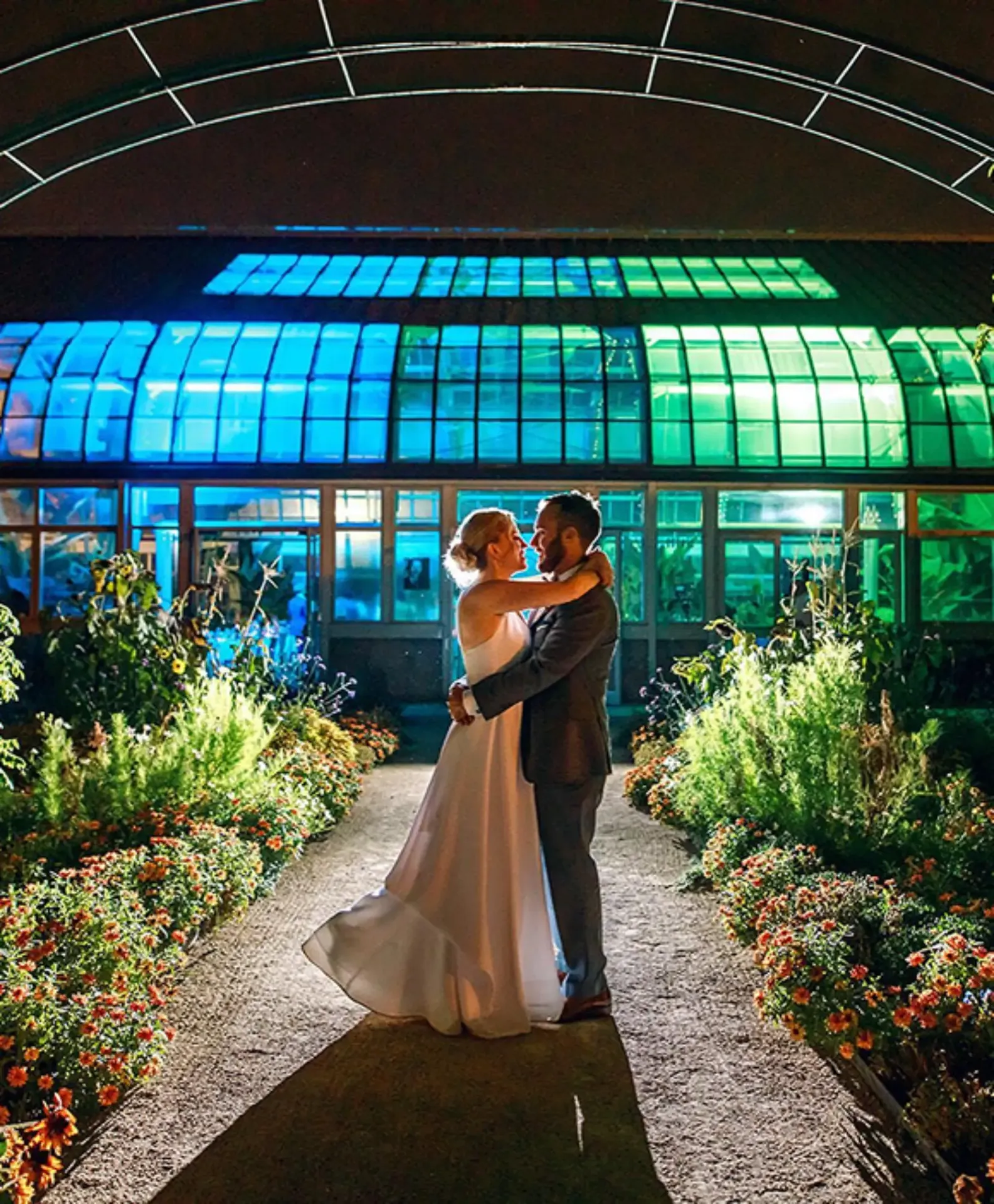 Bride and groom embracing outside illuminated conservatory at night, surrounded by flower beds