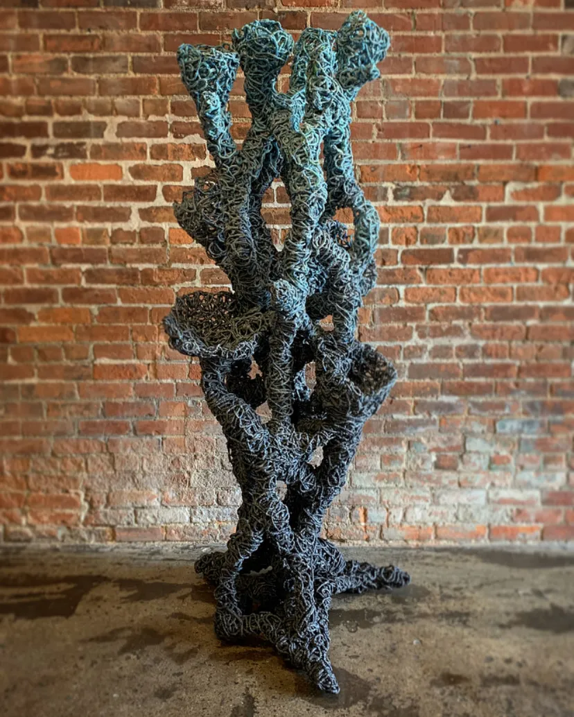 Plasticus Canistraria, a coral esque piece designed by Cody Norman stands against a brick wall. The scultpure is various shades of blue.