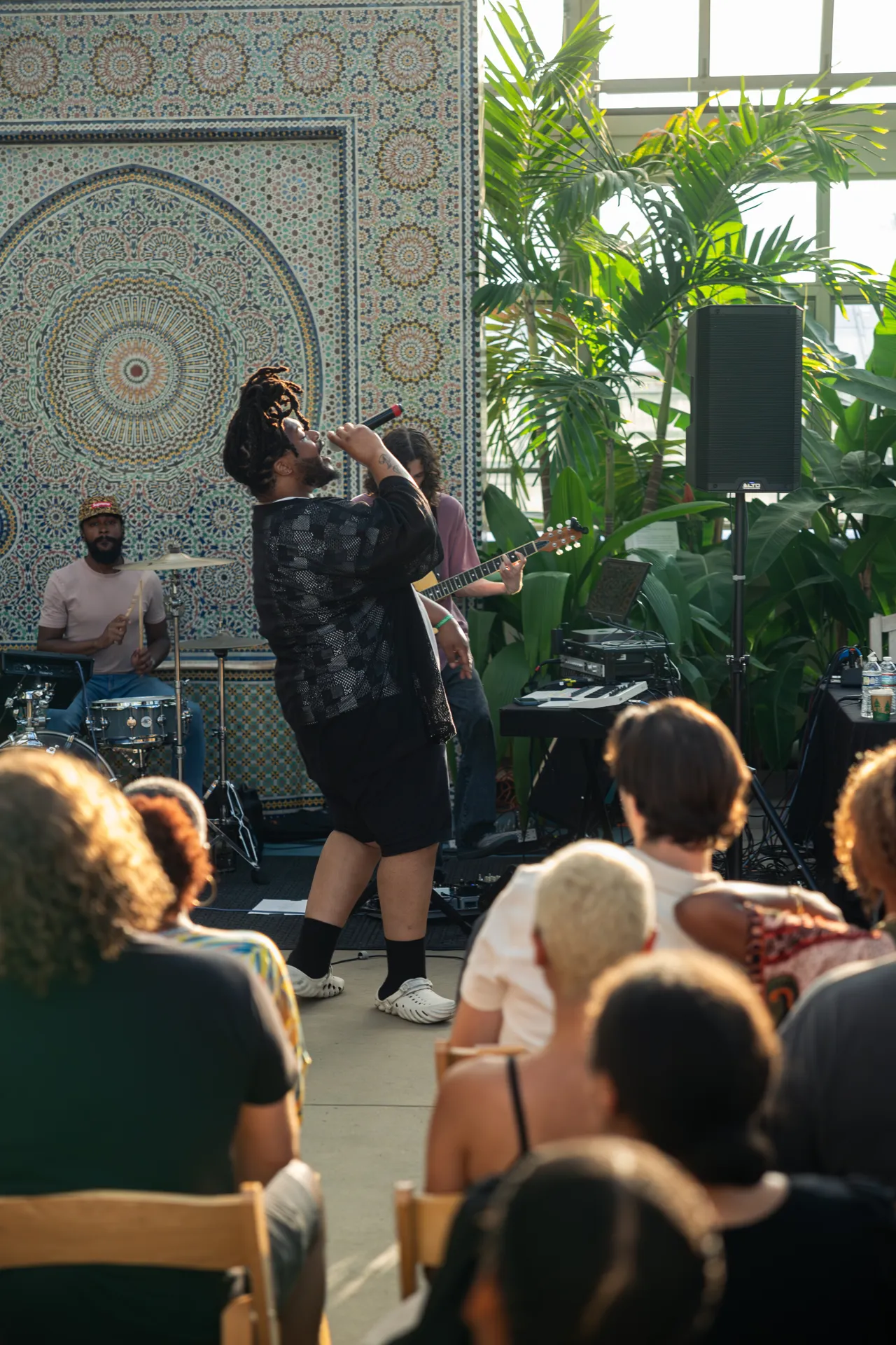 MfNMelo in the center of the photo singing into the microphone with the audience in chairs in the foreground.