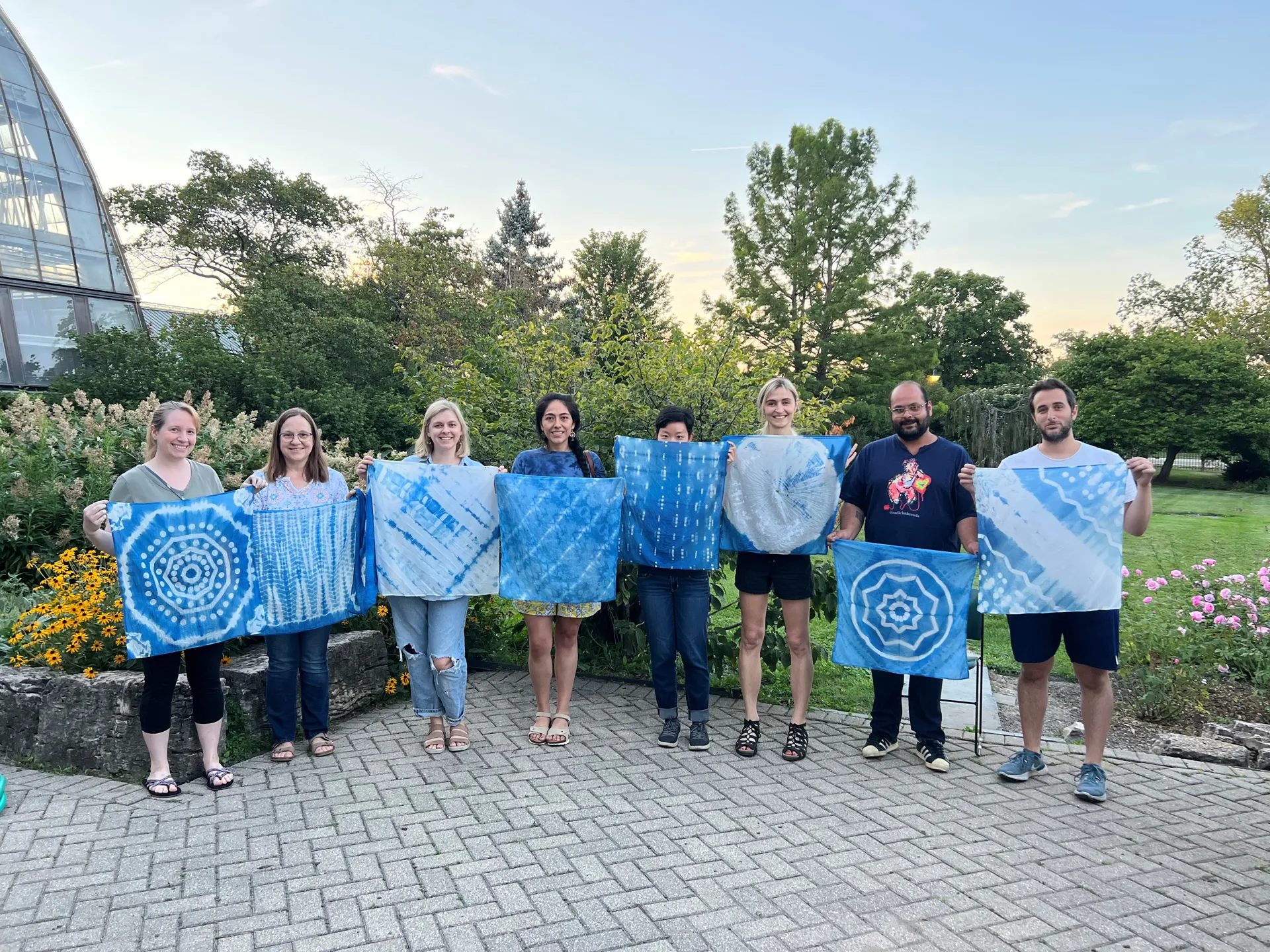 Eight people standing outside in a garden. Each person is holding up a bandana dyed with blue indigo in various unique patterns.