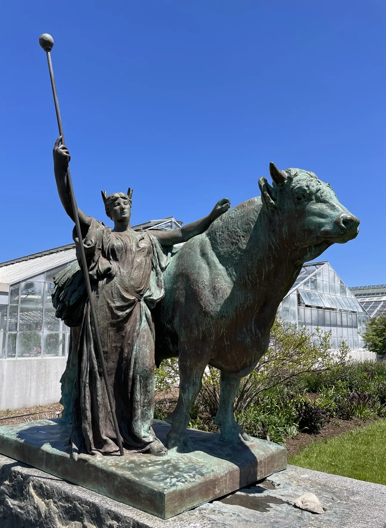 Bronze statue of a bull and maiden with greenhouses behind.
