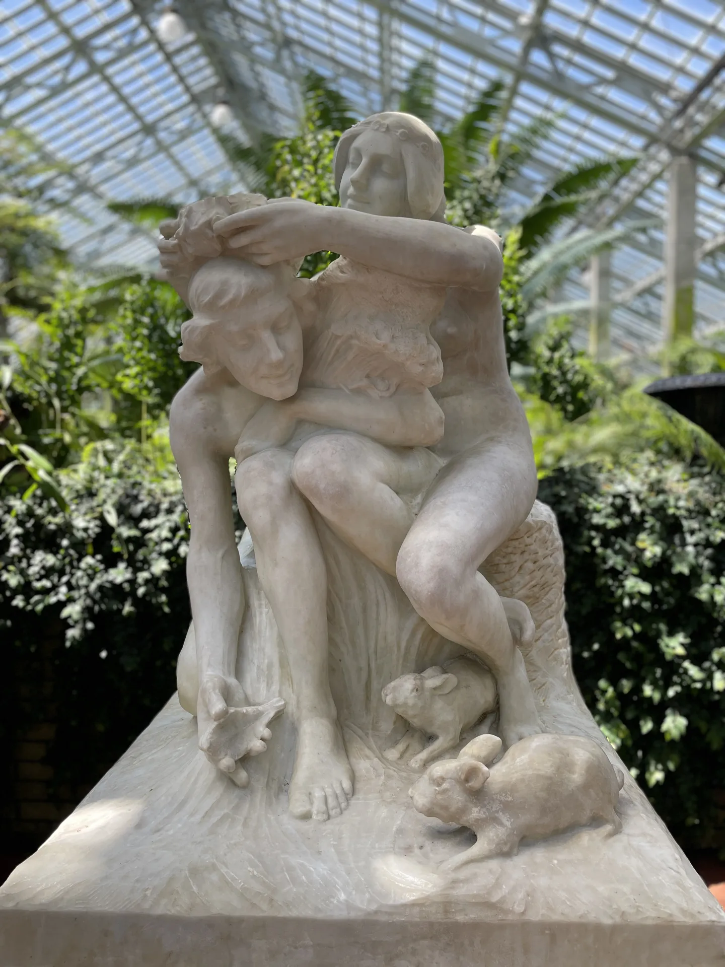 Off-white sculpture of man and woman in front of green plants.