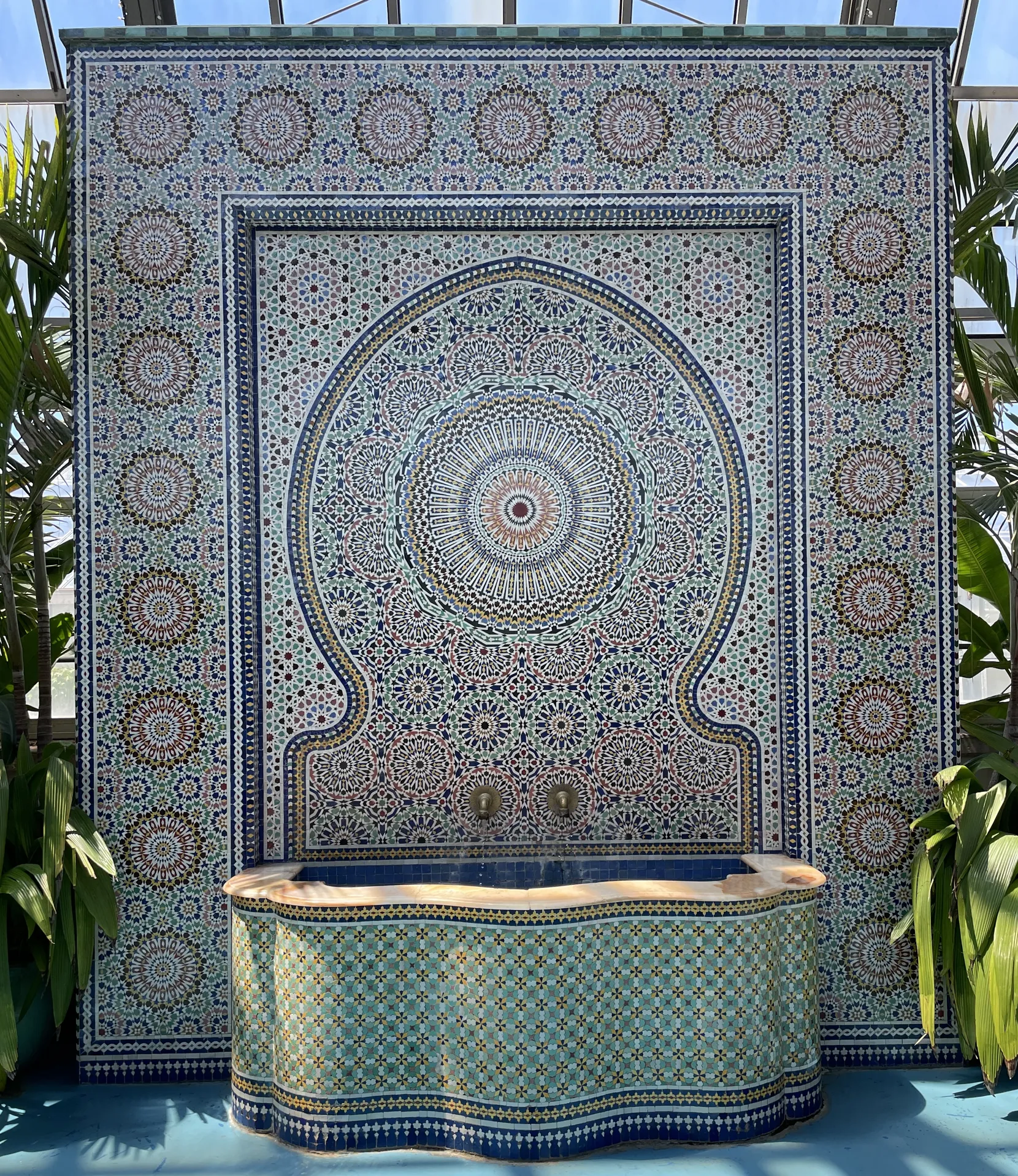 Zelijj or Moroccan Fountain with plants surrounding. The mosaic fountain features a water basin at the bottom middle.