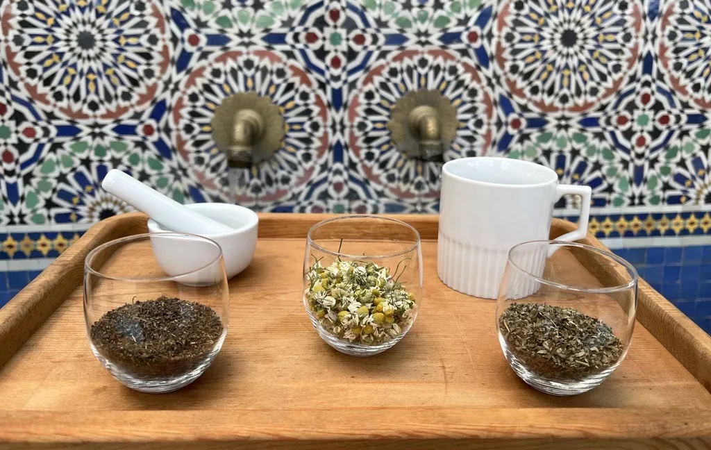 Three glass cups with dried herbs, a white mortar and pestle, and a white coffee mug on a wooden tray. Background: a mosaic fountain.