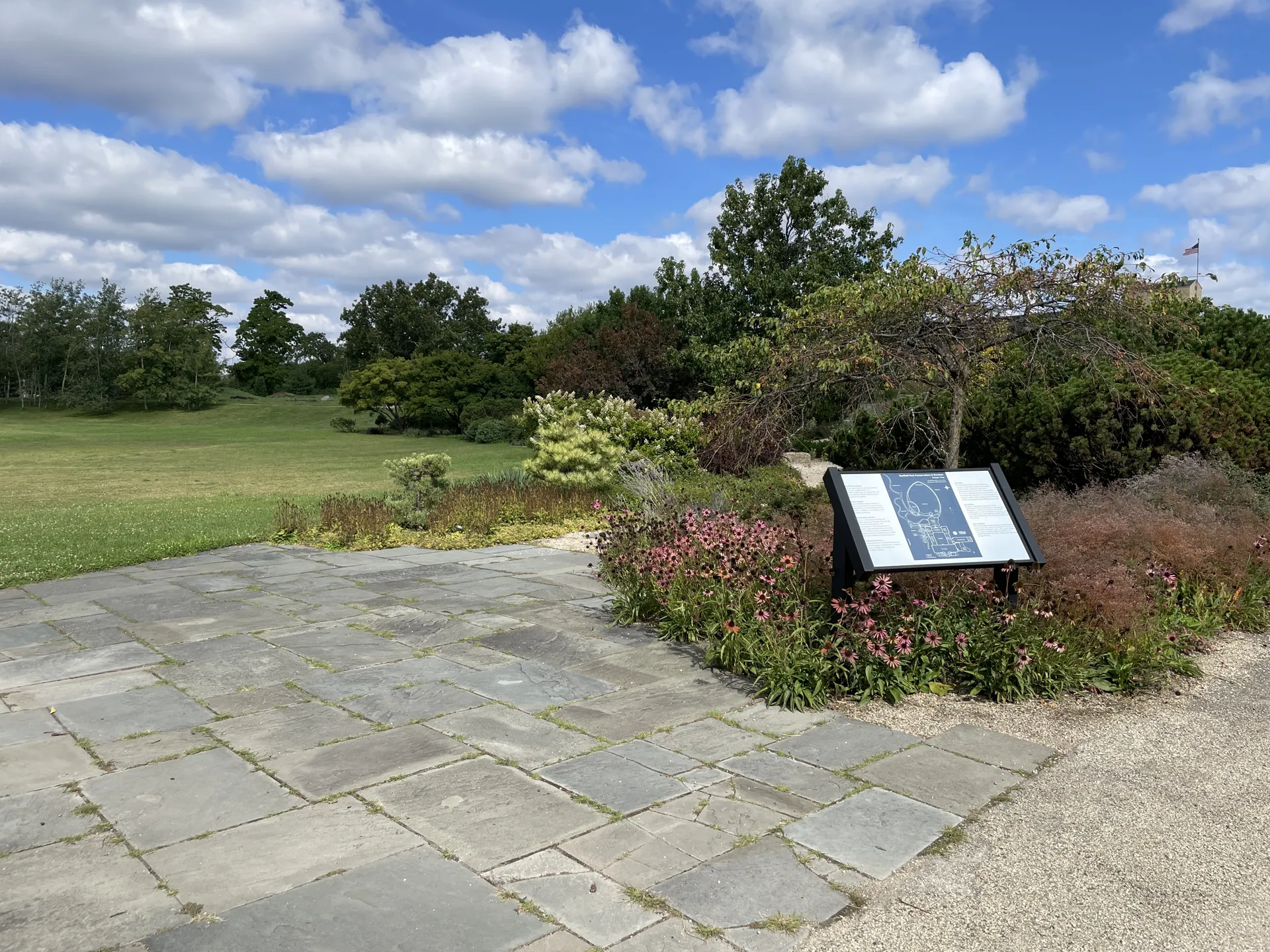 Outdoor garden view to the north west from the edge of the Bluestone Patio. A sign with a map, paved pathway and bluestone pathway in the foreground, grass and trees in the distance.