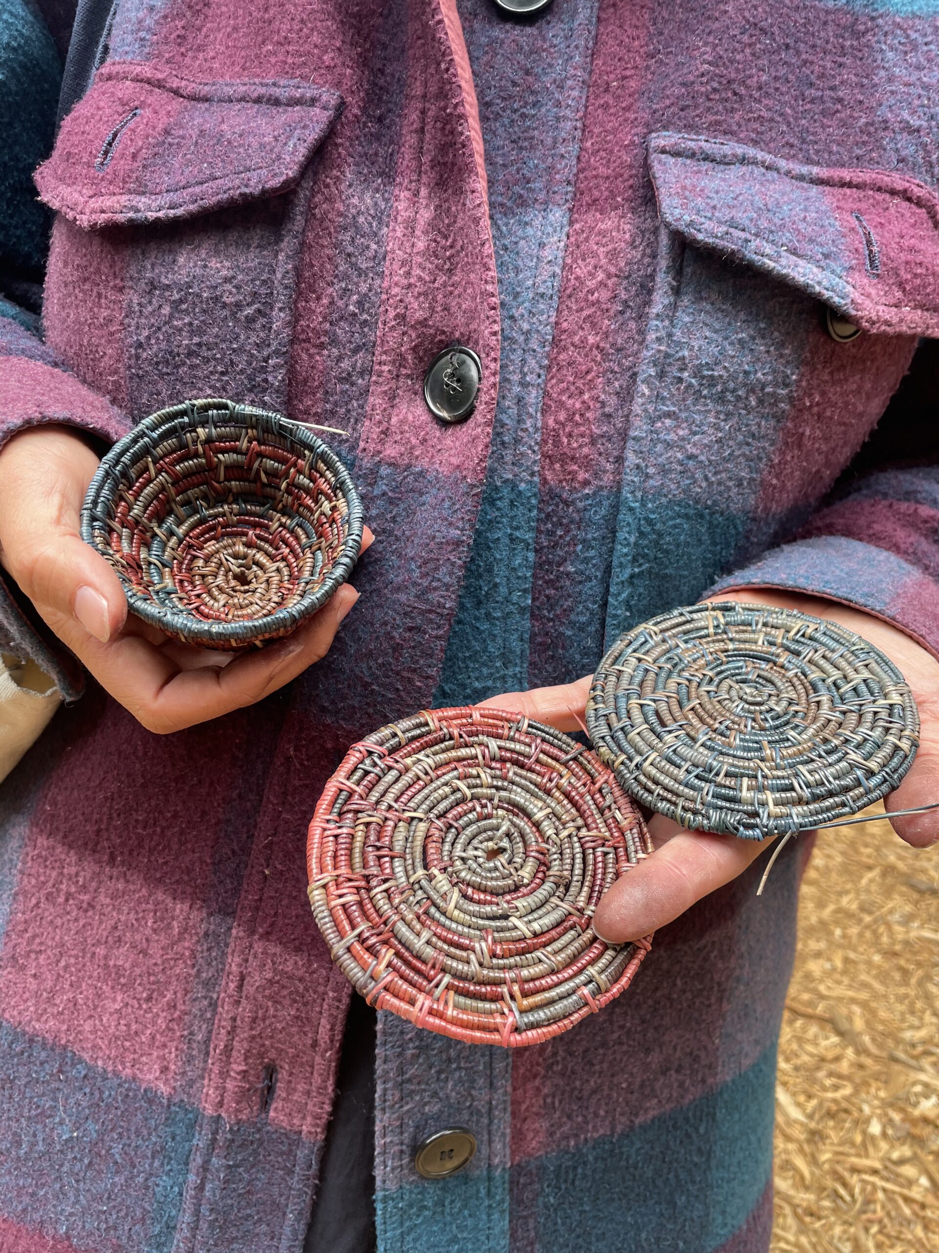 a woman in a purple plaid jacket holding three in =progress pine needle baskets.