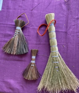 Three woven brooms on a purple back ground. Two are smaller made from pine needles. One is larger made from broom cord (sorghum)