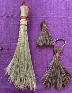 Three woven brooms on a purple back ground. Two are smaller made from pine needles. One is larger made from broom cord (sorghum)